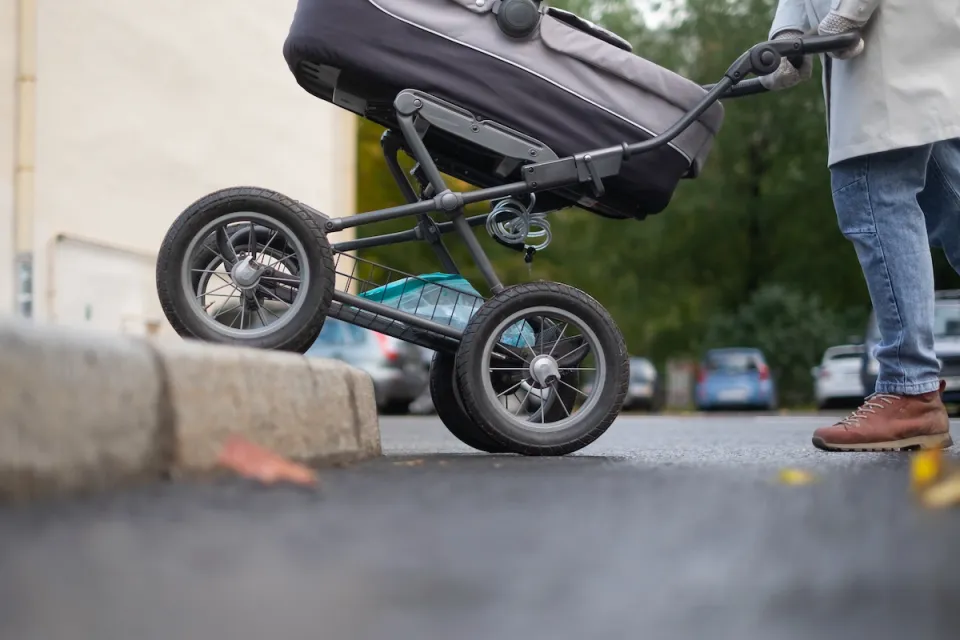 Close-up of bottom half of stroller being pushed onto sidewalk with no curb cut by person in jeans and brown shoes.